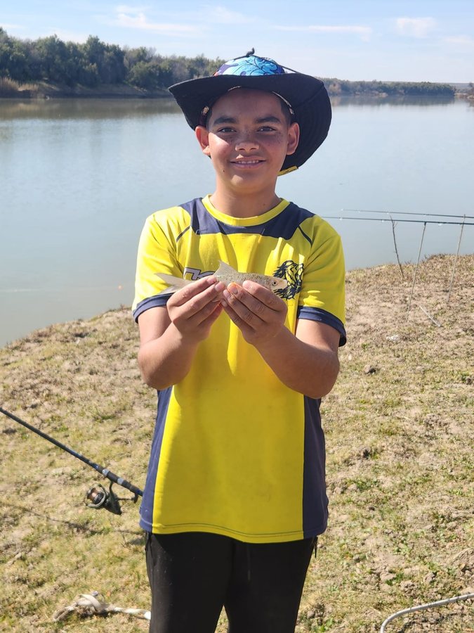 A young angler in a wide-brimmed hat holding a small fish by the riverbank, with a fishing rod resting nearby.