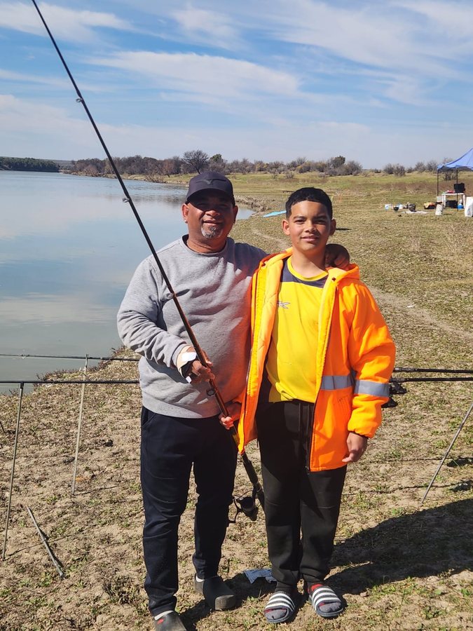 A father and son standing together by the water's edge, arm around each other's shoulders.
