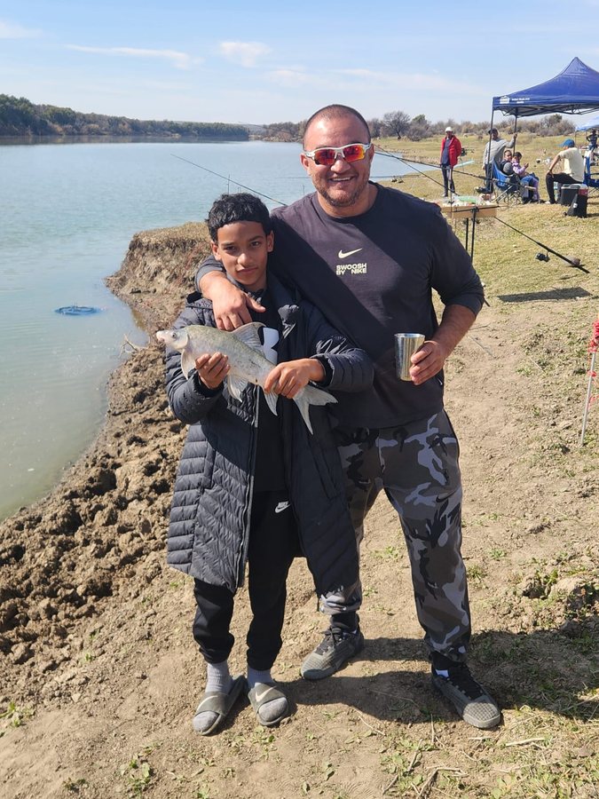 A father with his arm around his son, who holds a freshly caught fish, at a riverside fishing event.